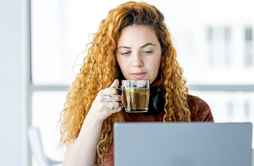 A red haired young woman takes a break using her laptop while drinking her hot tea.