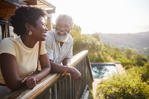 Couple laughing while standing on the balcony of their holiday rental and looking at the scenic view during a vacation