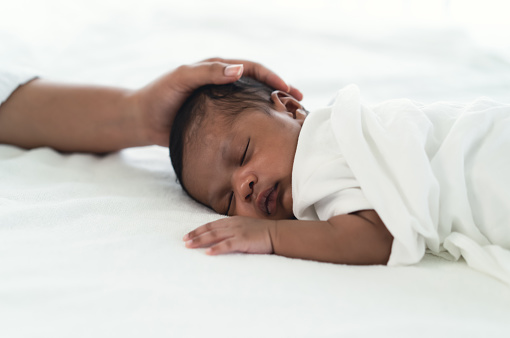 Portrait of Infant Black African American sleeping on white bed with mother hand in bedroom. During Daytime Sleep At Home. Newborn baby health care concept.