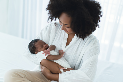 Beautiful African american mother holding lean to chest cute little infant toddler in her arms. Black mom looking baby with love in bedroom. Enjoying motherhood, soft focus, happy black family concept