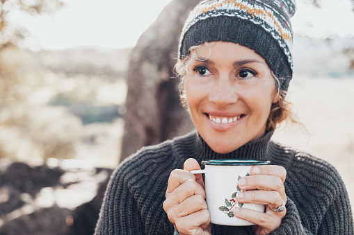 Face portrait of cheerful middle age pretty woman drinking cup of tea or coffee in outdoor leisure activity alone. Smiling lady looking on side. Nature outdoors background. Female people with knit hat