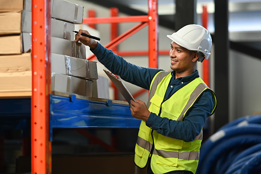 Smiling man warehouse workers with safety hard hat and vest checking quantity of storage product on shelf.