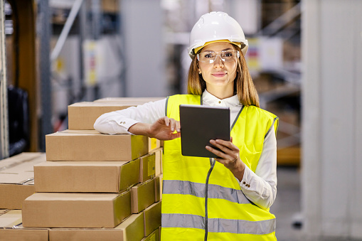 An inventory manager in work wear is using tablet in warehouse and looking at the camera.