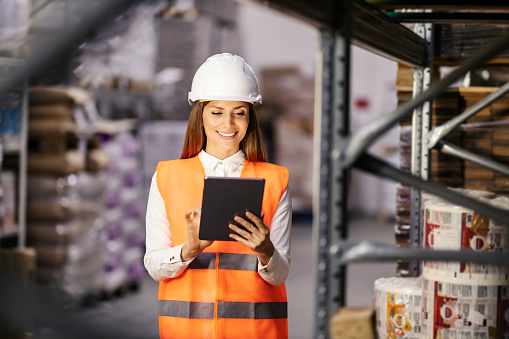 A warehouse manager is standing in facility and doing logistics on tablet for wares.