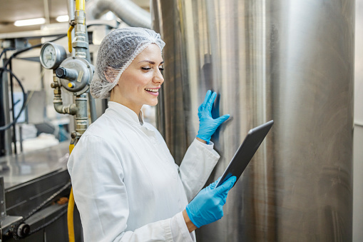 A milk processing factory worker is checking on machine with milk while holding tablet and smiling at it.