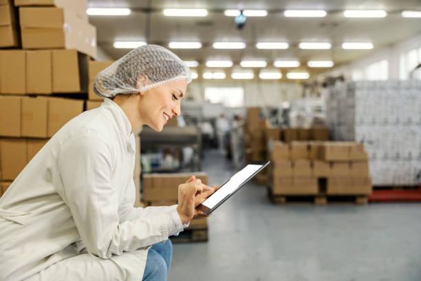 A food factory manager is crouching and scrolling on tablet in facility. stock photo