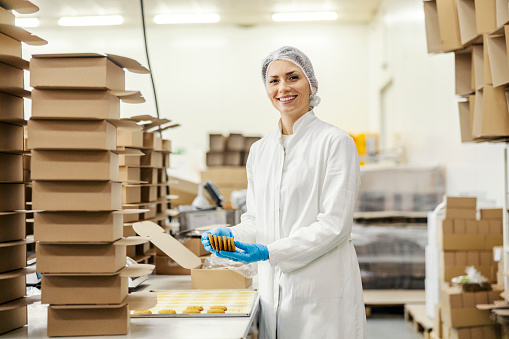 A food plant worker is collecting cookies while smiling at the camera.