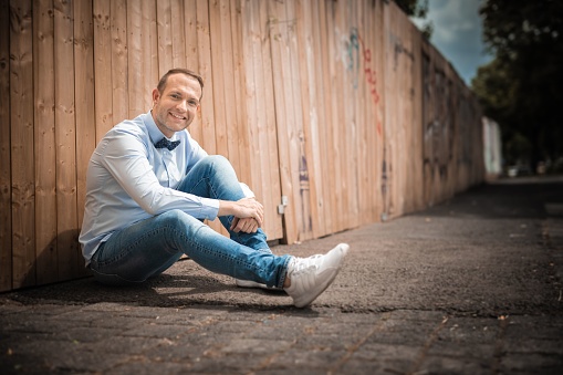 An adult man sitting on the ground in front of a wooden fence and expressing positive emotions