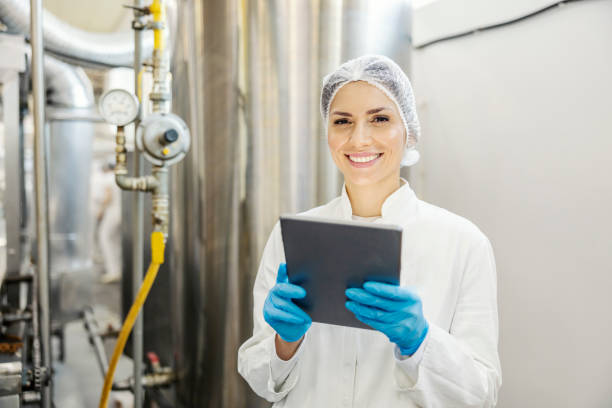 un ispettore di una fabbrica di latte è in piedi accanto a una macchina per la lavorazione del latte con il tablet in mano e sorride alla telecamera. - stabilimento caseario foto e immagini stock