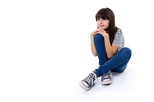 A young 10 year old girl sit on floor rest with hands on knee and smile into camera with copy space isolated on white background