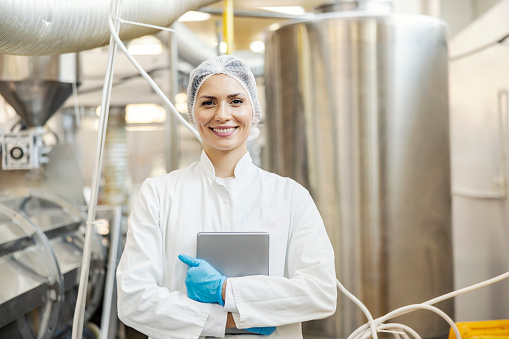 A dairy factory worker is standing in facility and holding tablet while smiling at the camera.