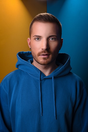 A vertical of a serious man in a hoodie looking into a camera on a blue-yellow background