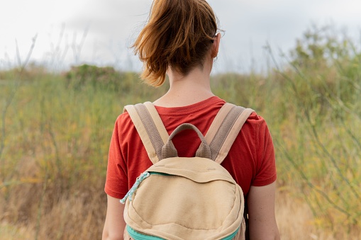 Back view of a woman in an orange t-shirt with a backpack walking in nature, freedom, travel and wellbeing concepts