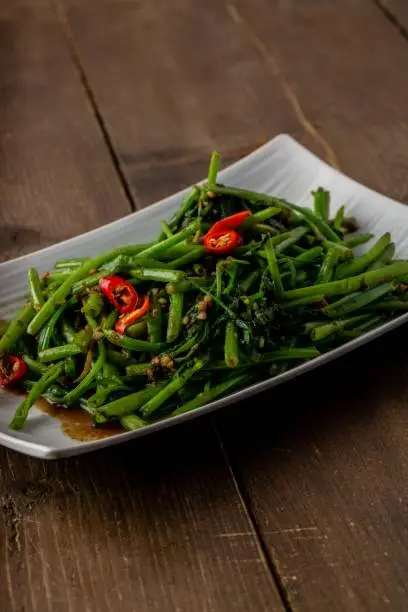 A closeup shot of a Chinese Sambal Kangkong dish, water spinach stir-fried with a spicy sauce, and sliced chili pepper, served on a white plate