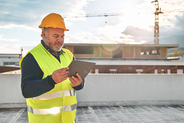 istock Middle aged smiled bearded supervisor in hardhat and safety vest making notes in tablet on building site. Structural engineer or architect monitors progress of work on construction site. 1453066783