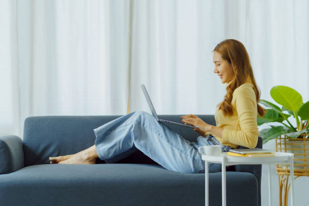 istock Smiling young Asian woman using laptop sitting on sofa at home 1453069550