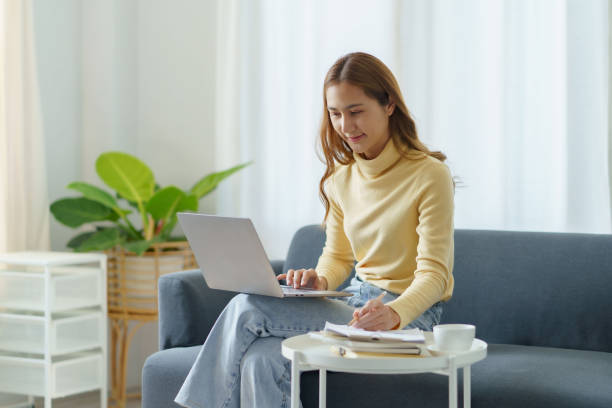 istock Smiling young Asian woman using laptop sitting on sofa at home 1453069551