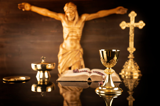 Catholic symbols composition: cruciefied Jesus figure, The Cross, monstrance,  Holy Bible, rosary and golden chalice on brown background.