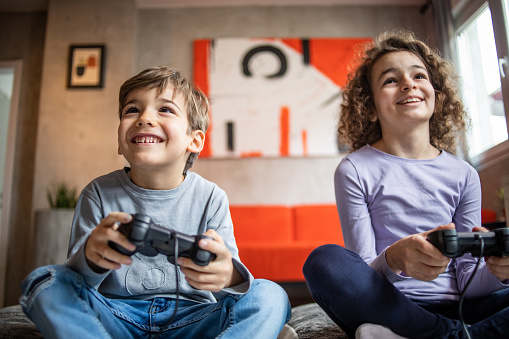 Two kids, brother and sister play video games using joystick while sitting at home on living room floor, use free time to have fun with video games