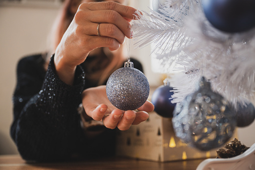 Closeup view of female hands about to hang a shiny blue holiday bauble on white christmas tree in a domestic life image.