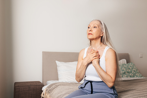 Worried senior woman sitting on a bed. Sad adult female with hands on chest.