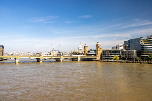 Cannon Street Railway Bridge. In the background is the skyline of the City of London including global architectural buildings and people are visible on the riverbank.