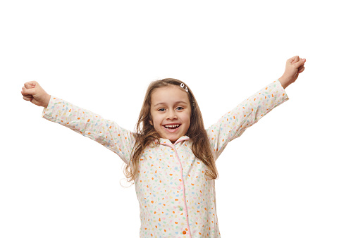 Cheerful baby girl in white pyjama with colorful dots, raising arms up, rejoicing, expressing amazement and happiness, smiling looking at camera, on white background. Space for your advertising text
