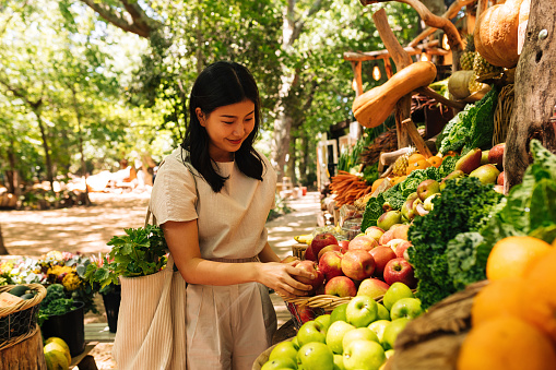Smiling Asian woman choosing fruits. Female with a shopping bag at an outdoor market.
