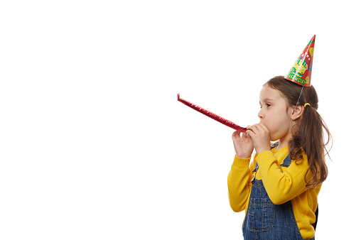 Side portrait of a charming Caucasian child, lovely birthday girl, dressed in yellow pullover, blue denim overalls and birthday hat, blowing in whistle on white background. Copy advertising space