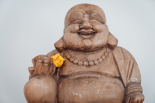 A statue of a smiling Buddha with a yellow flower on a white wall background