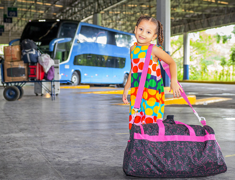 Little Brazilian girl, black or brunette, with an afro hairstyle, wearing a multicolored dress and a large travel bag waiting for the bus at the bus station