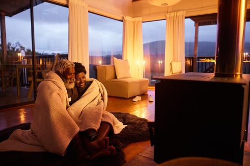 Affectionate couple sitting together wrapped in a blanket in front of a fireplace in there living room with a view