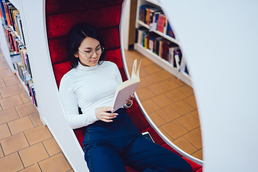 High angle of concentrated serene ethnic woman in eyeglasses having break on stylish bench in modern library while reading book