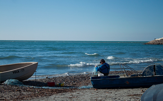 A fisherman untangles his fishing net while seated on his small fishing boat in a warm winter December afternoon in the coastal city of Durres, Albania. The Adriatic Sea is calm.