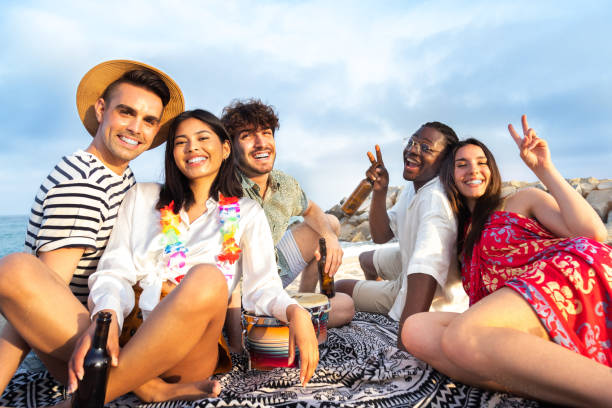 szczęśliwa, uśmiechnięta grupa wielorasowych przyjaciół relaksujących się przy piwie, pijących razem i patrzących w kamerę. - party beach indian ethnicity adult zdjęcia i obrazy z banku zdjęć