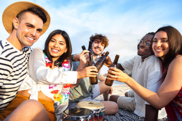 szczęśliwa wielorasowa grupa przyjaciół wznoszących toasty piwem na pikniku na plaży, patrząc w kamerę. - party beach indian ethnicity adult zdjęcia i obrazy z banku zdjęć