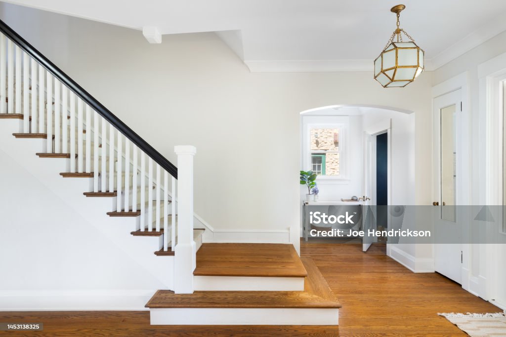 A home's foyer with a renovated staircase and gold light fixture hanging above. A renovated stairway in the home's foyer with a view of a mud room and doorway to a small, blue bathroom. A modern light hangs above the hardwood floors. Staircase Stock Photo