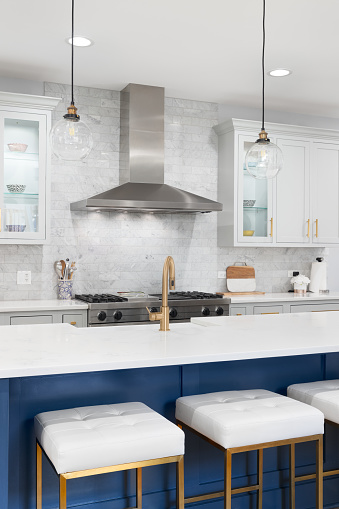 A beautiful modern kitchen with grey and blue cabinets, stainless steel appliances, gold hardware and glass lights hanging above the large island.
