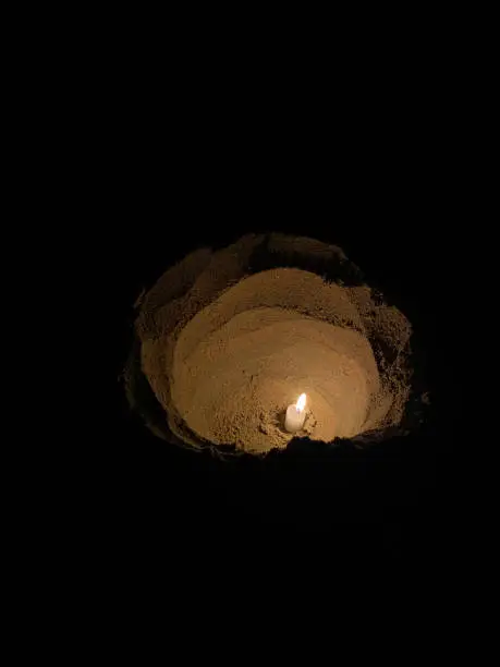 Photo of Image of individual, wax candle in deep hole dug in beach sand, lit wick, flickering flame glowing in dark, sandy beach at night, elevated view, focus on foreground