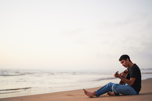 young latino male playing guitar and singing alone relaxed sitting on the beach shore at sunset