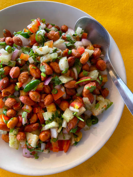 Stock photo showing elevated view of homemade peanut masala chaat recipe made with peanuts, chaat masala, chilli powder, salt, lemon juice, coriander, sliced serrano pepper (Capsicum annuum), diced tomato and red onion, served in a white bowl, with a metal spoon. imagen de tazón blanco con cuchara de metal que contiene masala chaat de maní casero, bocadillo salado al estilo callejero, cacahuetes, chaat masala, chile en polvo, sal, jugo de limón cilantro, chile serrano en rodajas (capsicum annuum), tomate cortado