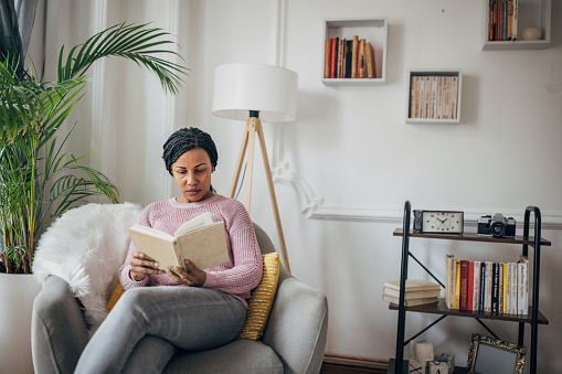 One woman, black woman sitting in armchair and reading a book in her home.