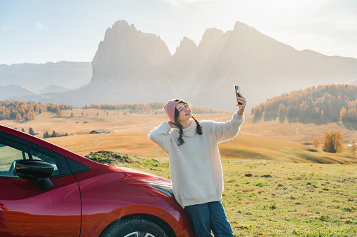 Young Caucasian woman  standing near the car on the background of Seiser Alm in Dolomites, Italy  and taking selfie with smartphone