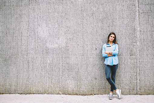 Full length portrait of attractive female millennial with electronic touch pad in hands looking at camera standing at promotional urbanity, Caucasian hipster girl with digital tablet posing in city