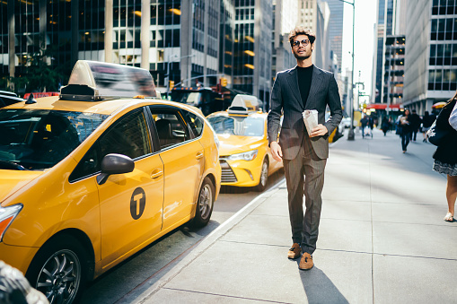 Serious male manager 30s dressed in elegant trendy wear and eyeglasses spending time outdoors, handsome businessman in formal suit walking near traffic road with taxi in metropolitan downtown