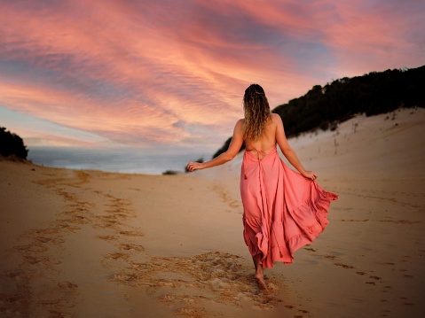 A back view of a young woman in a flowy dress walking on a sandy beach against a beautiful pink sunset