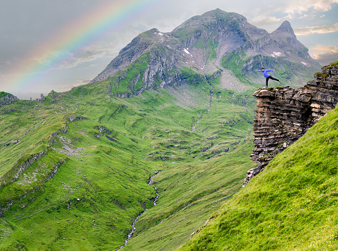 Mature woman standing acrobatically on the edge of cliff in the Swiss alps near Grindelwald with rainbow in background.