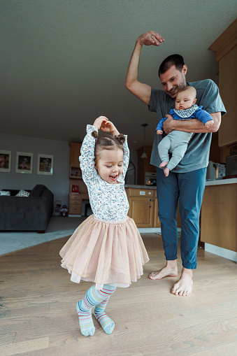A playful dad affectionately holds his four month old baby and dances with his cute toddler daughter in the kitchen while spending time together at home.