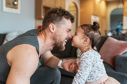 Profile view of a dad and toddler girl giggling and touching noses while playing together at home in the living room.