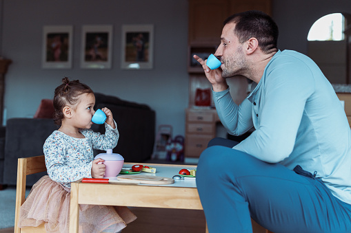 Profile view of a dad and his toddler daughter sitting at a child size table and pretending to sip tea from toy teacups while playing together at home.